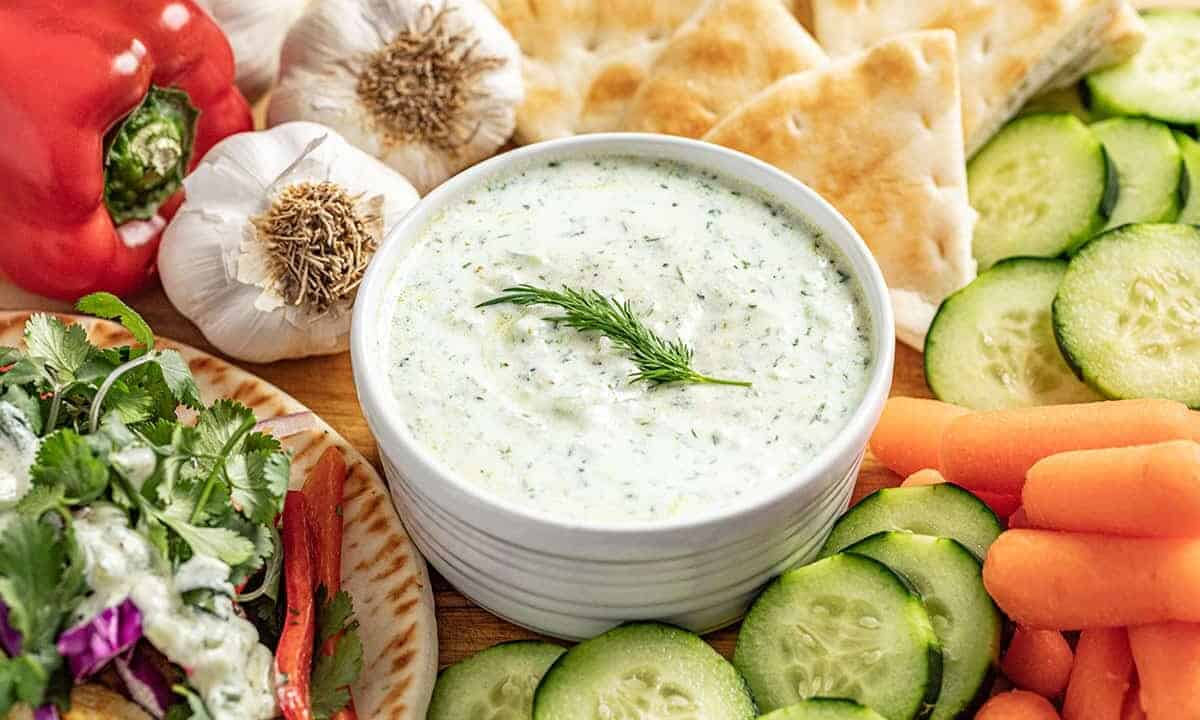 Tzatziki sauce in a white bowl topped with dill surrounded by various vegetables.