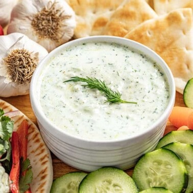 Tzatziki sauce in a white bowl topped with dill surrounded by various vegetables.