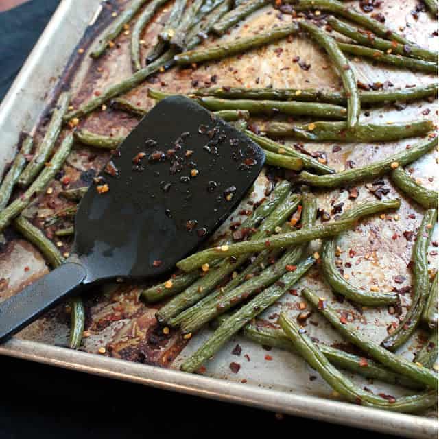 Black spatula resting on a sheet pan with roasted green beans garnished with spicy red pepper flakes