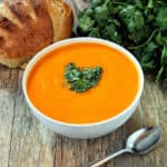 Bright orange soup in a white bowl garnished with parsley and a loaf of bread in the background