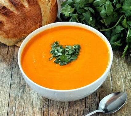 Bright orange soup in a white bowl garnished with parsley and a loaf of bread in the background