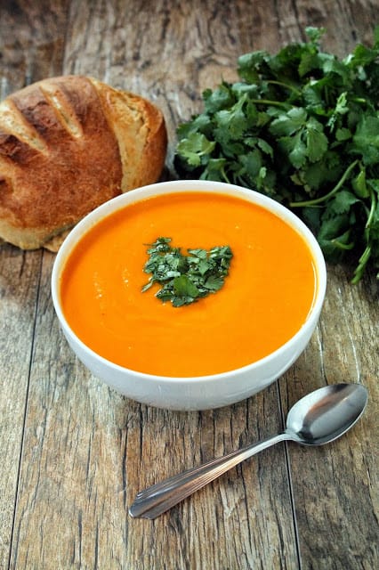 Bright orange soup in a white bowl garnished with parsley and a loaf of bread in the background
