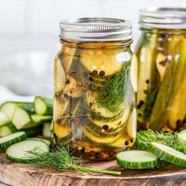 2 jars of refrigerator pickles on a serving board with fresh cucumbers, dill, and garlic close by