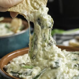 pulling out a tortilla chip dipped in spinach artichoke dip with melted cheese stretching down to the bowl