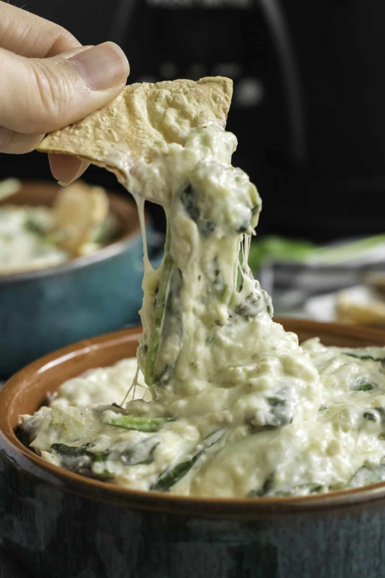 pulling out a tortilla chip dipped in spinach artichoke dip with melted cheese stretching down to the bowl