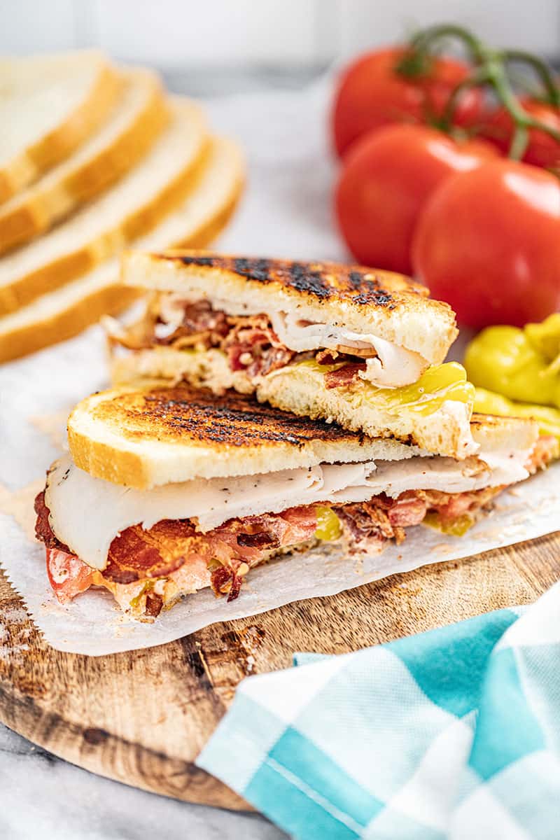 baja turkey club sandwich cut in half on a wooden board with fresh tomatoes in background