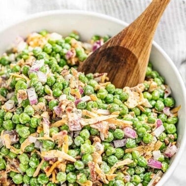 A white bowl filled with pea salad with a large wooden spoon inside