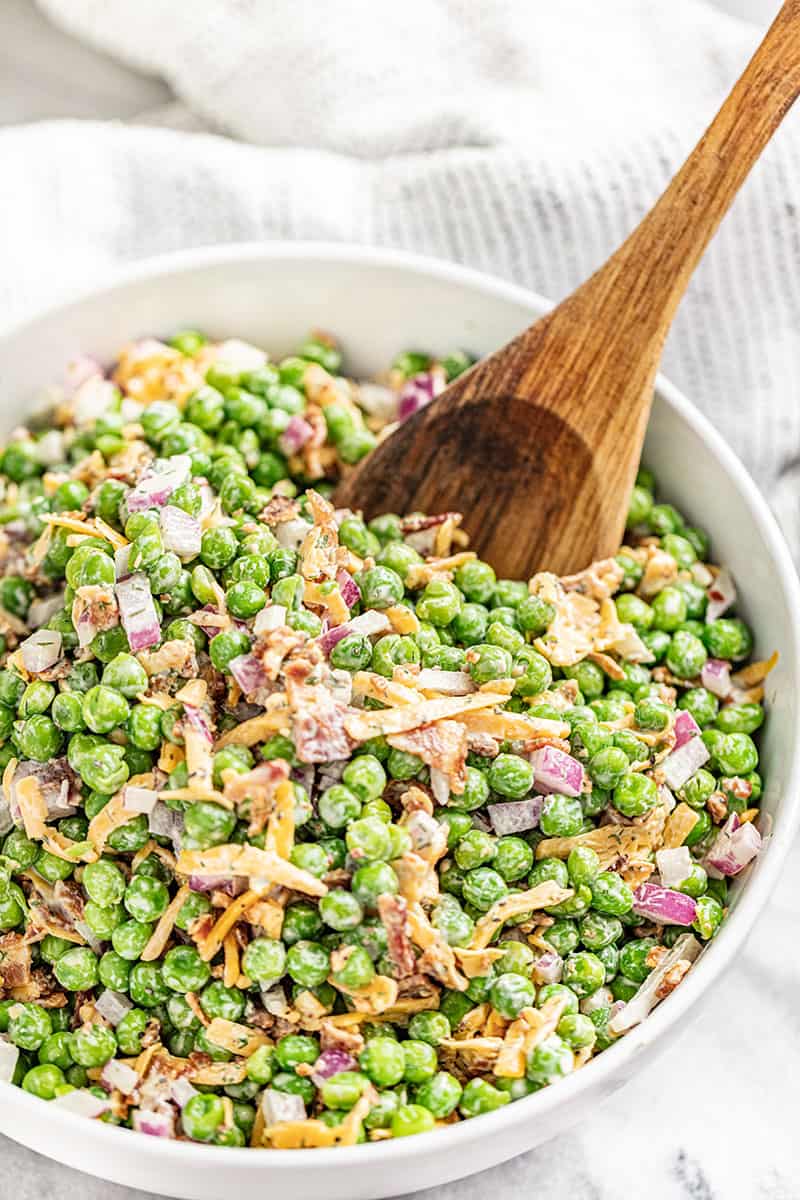 A white bowl filled with pea salad with a large wooden spoon inside