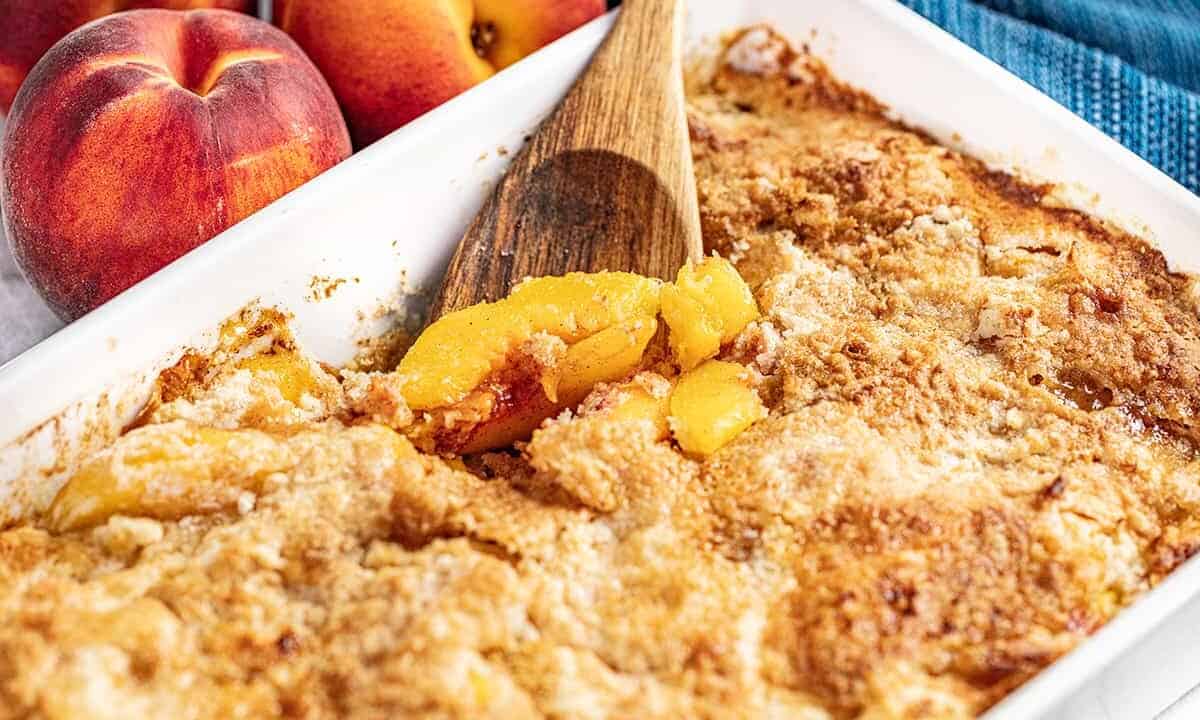 A baking pan full of peach dump cake with a wooden spoon digging in