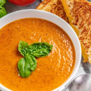 tomato basil soup with basil on top in a white bowl with a grilled cheese nearby