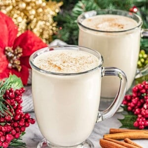 2 glass mugs of homemade eggnog with a Christmas tree and holly and ivy in the background
