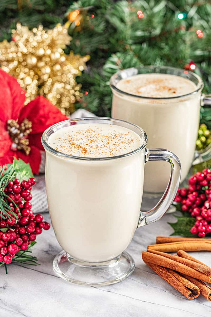 2 glass mugs of homemade eggnog with a Christmas tree and holly and ivy in the background.