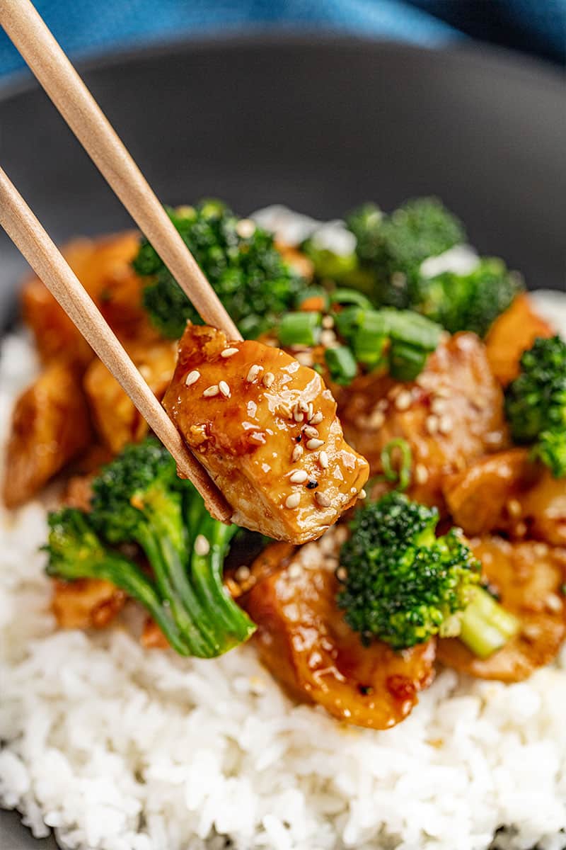 Chopsticks holding a piece of chicken teriyaki covered in sesame seeds.