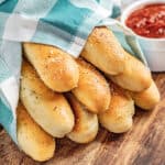 Homemade breadsticks wrapped in a blue checkered towel with a small bowl of marinara sauce nearby.