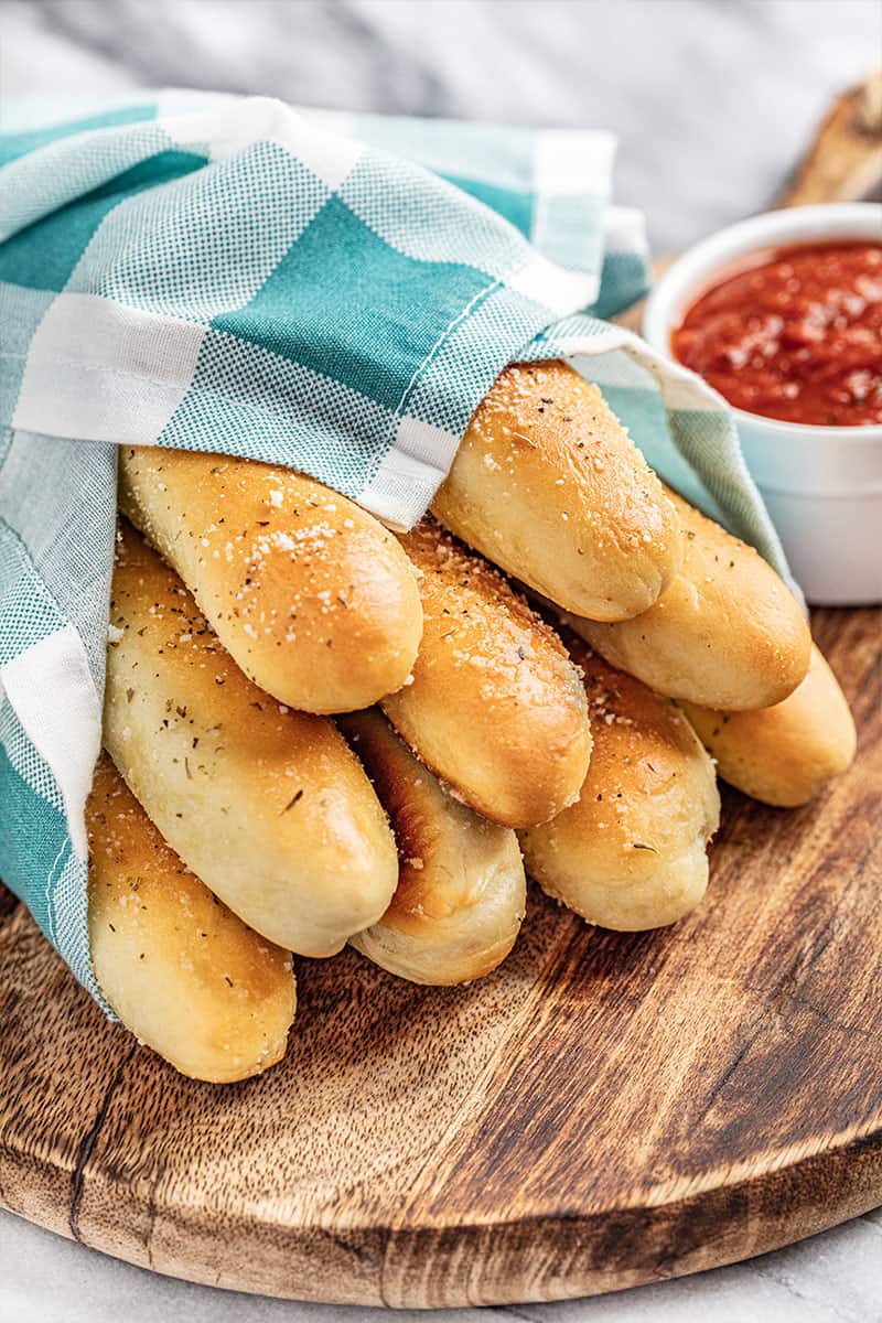 Homemade breadsticks wrapped in a blue checkered towel with a small bowl of marinara sauce nearby.