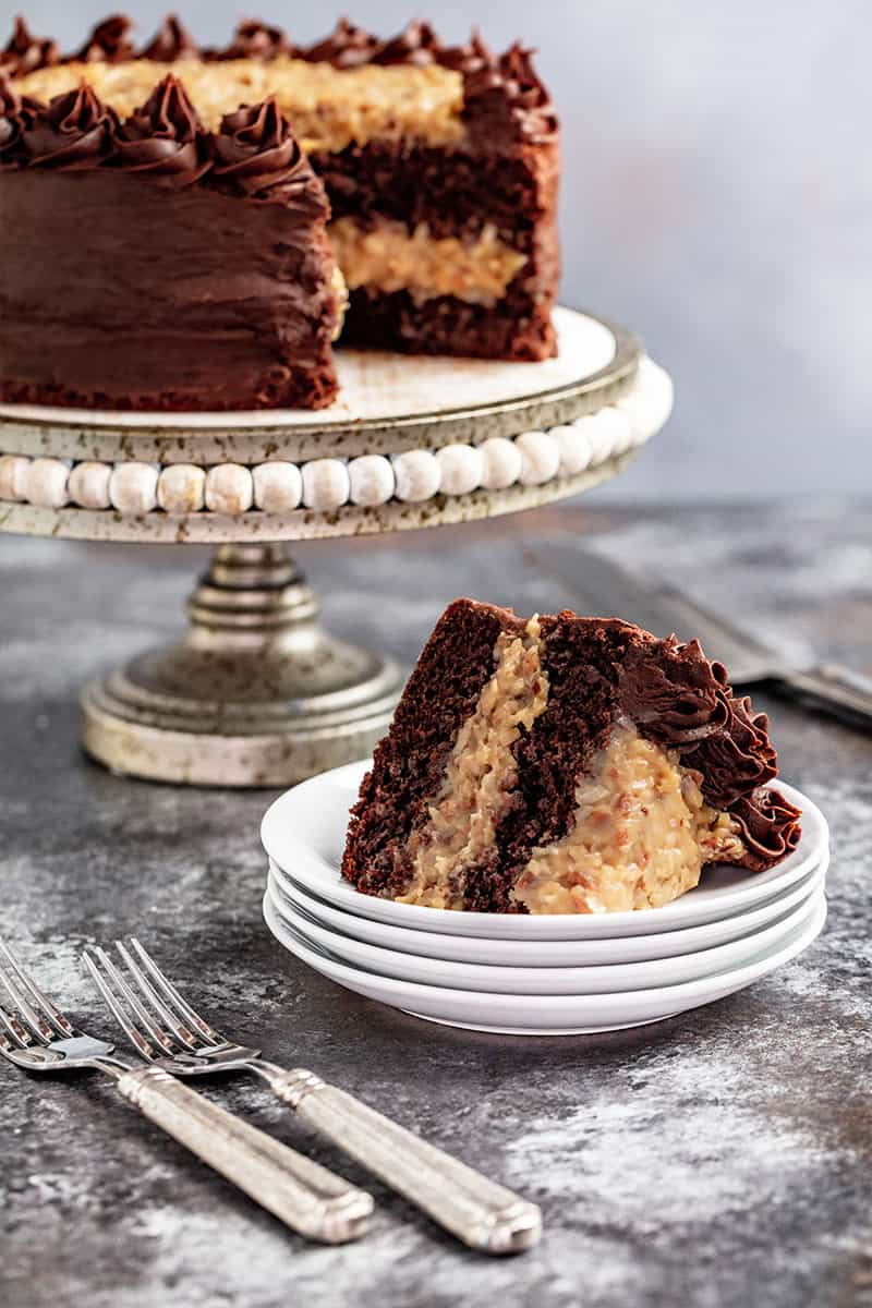 A slice of german chocolate cake on a stack of white plates with the whole cake on a stand in the background.