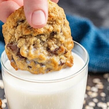 A hand dunking an oatmeal chocolate chip cookie into a glass of milk.
