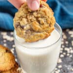 A hand dunking an oatmeal chocolate chip cookie in a glass of milk.