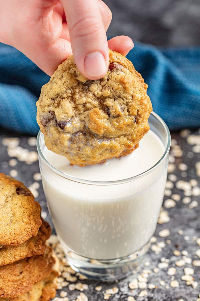 A hand dunking an oatmeal chocolate chip cookie in a glass of milk.