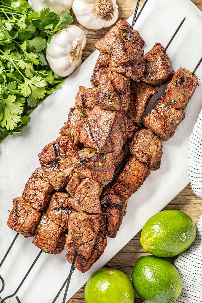 Overhead view of beef kabobs next to limes, garlic, and coriander.