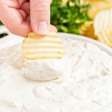 A hand dipping a crinkle cut potato chip into a serving dish filled with creamy French onion dip.
