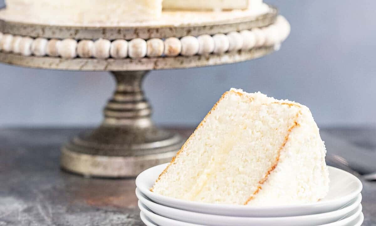 A slice of white cake on a stack of white plates with a whole white cake on a cake platter in the background.