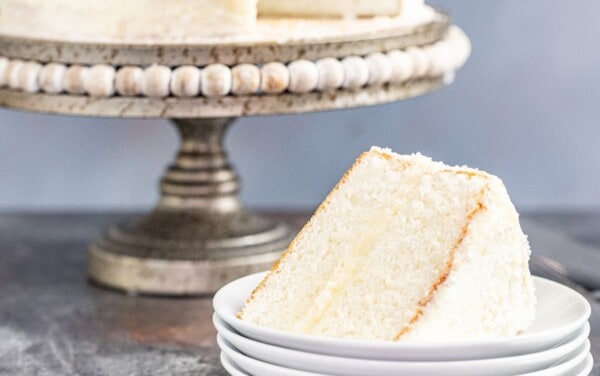 A slice of white cake on a stack of white plates with a whole white cake on a cake platter in the background.
