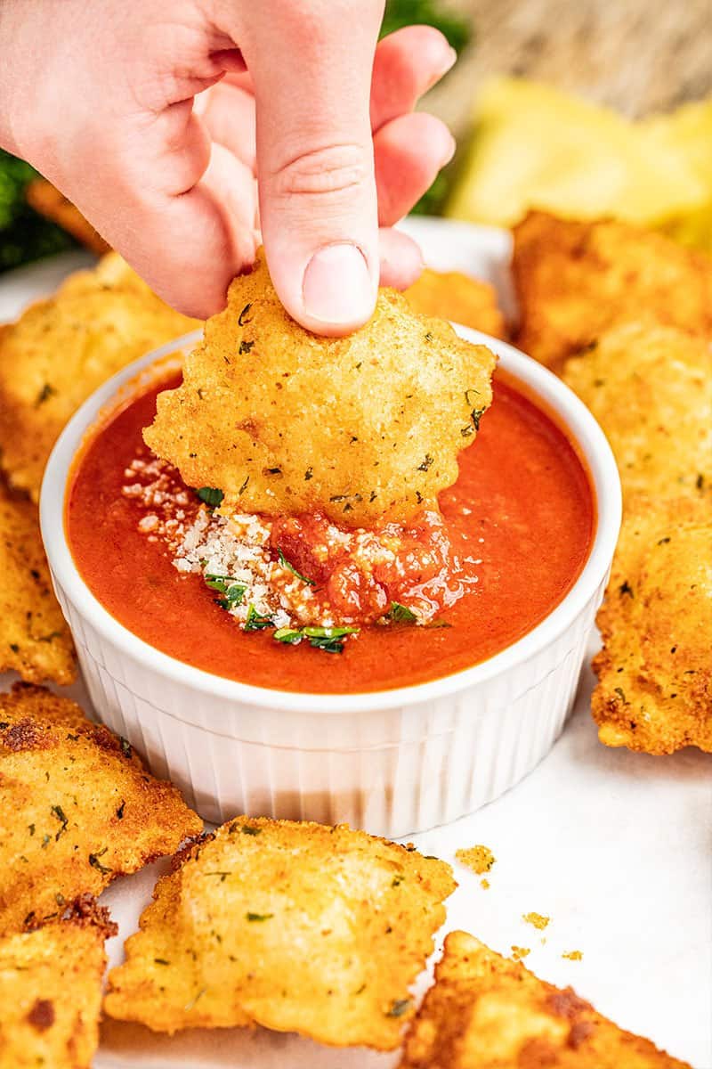 A hand dipping a fried ravioli into dipping sauce.