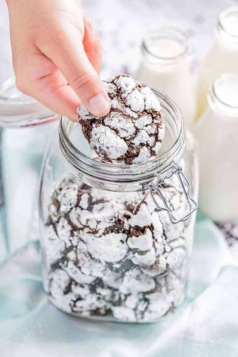 A hand holding a chocolate crinkle cookie over a glass jar filled with cookies.