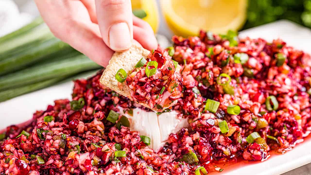 Close up view of a hand holding a cracker dipped in cranberry salsa.