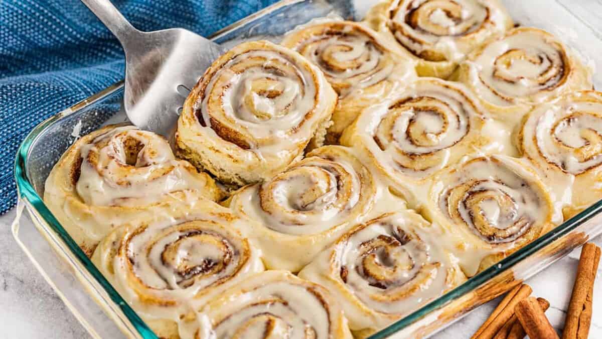 A spatula picking up a cinnamon roll from a glass baking pan.