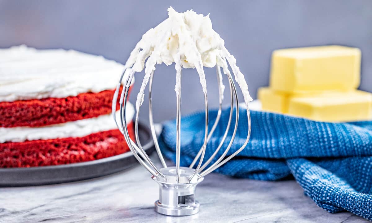 A stand mixer whisk attachment standing up with ermine frosting on it.