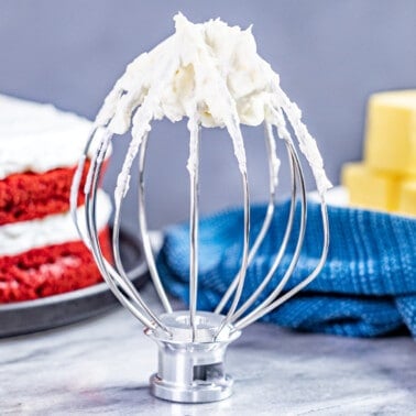 A stand mixer whisk attachment standing up with ermine frosting on it.