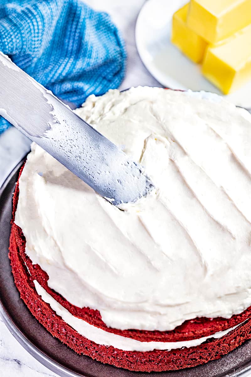 A knife spreading ermine frosting on top of a red velvet cake.