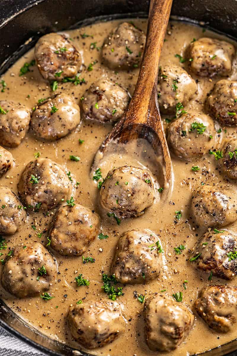Close up view of Swedish meatballs and gravy in a cast iron skillet with a wood spoon scooping up a meatball.
