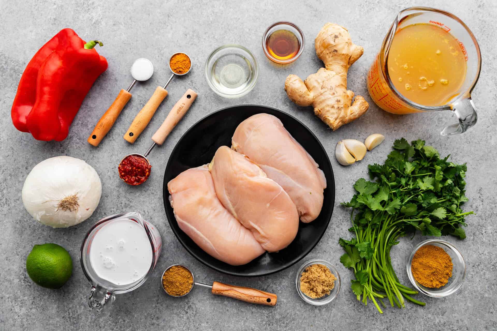 An overhead photo of all the ingredients needed to measure spoons and bowls around a plate of raw chicken and whole fresh vegetables.