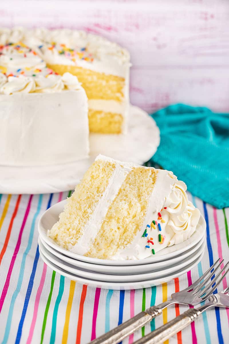 A piece of vanilla cake on a plate with the rest of the cake in the background.