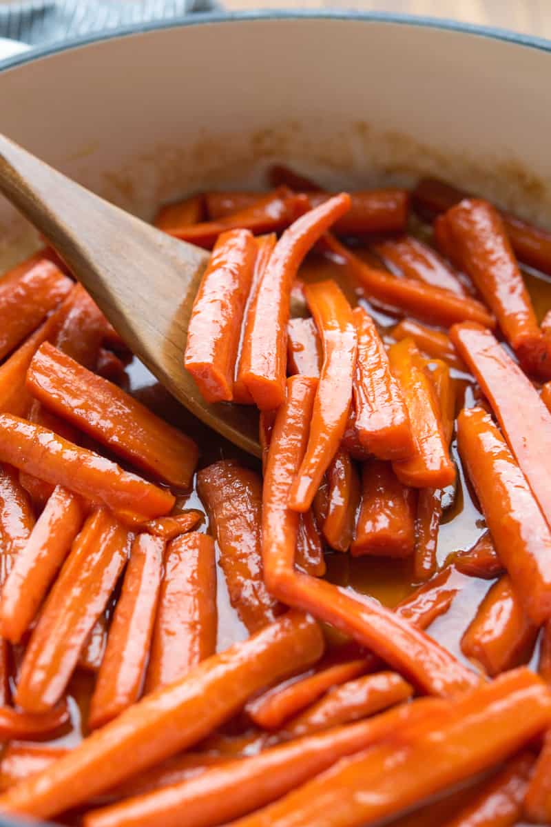 Close up view of a wooden spoon stirring glazed carrots in a pot.