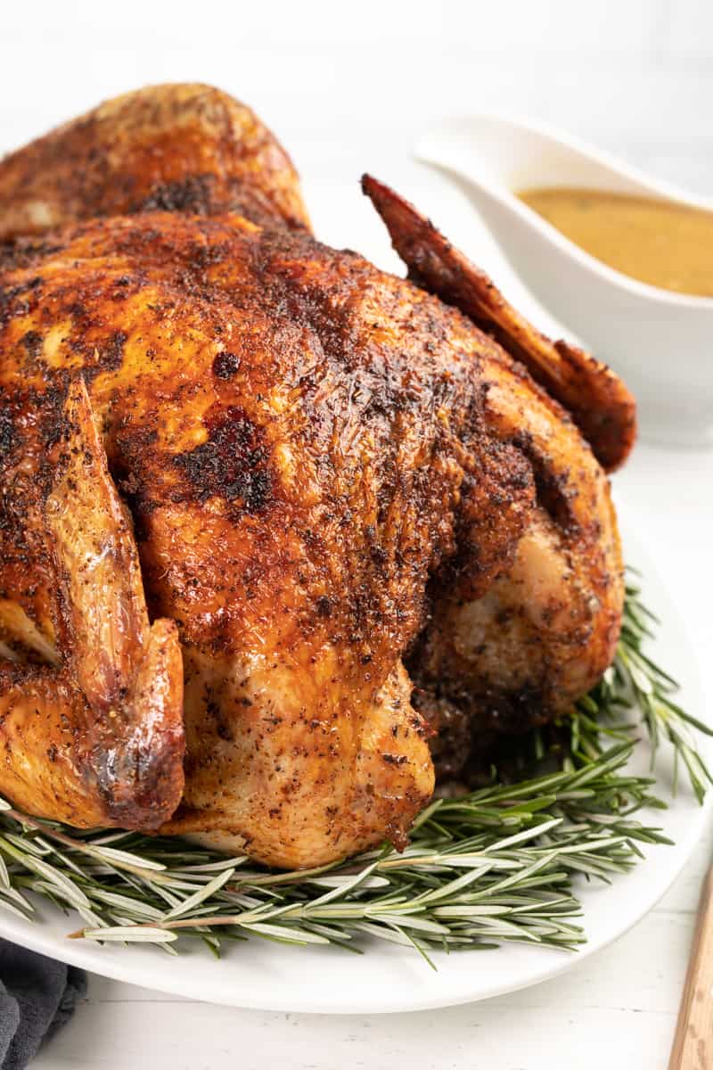 Large Thanksgiving turkey on a white platter with rosemary and a gravy boat in the background.