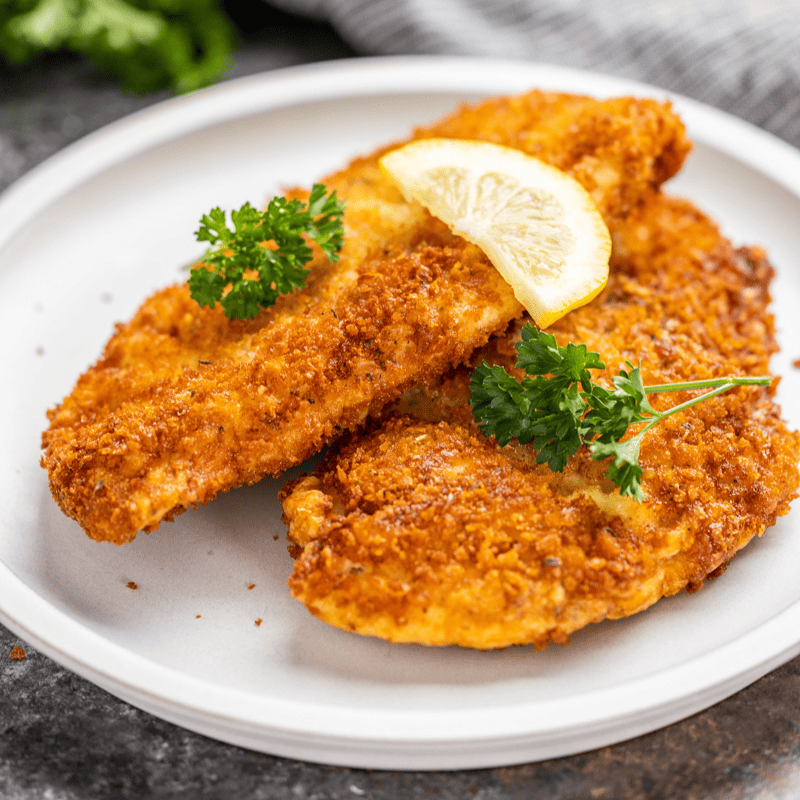 Two chicken cutlets on a white plate topped with a slice of lemon and some sprigs of fresh parsley.