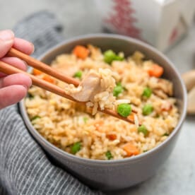 A hand holding chopsticks scooping out some chicken fried rice from a bowl.