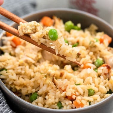 A hand holding chopsticks scooping out some chicken fried rice from a bowl.