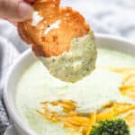 A slice of crusty toasted baguette being dipped into a bowl of cream of broccoli soup.