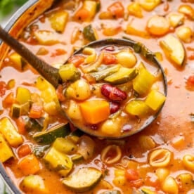 Overhead view of a white pot full of minestrone soup with a ladle full of soup being scooped out.