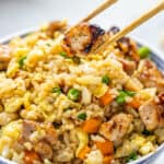 A bowl of pork fried rice with chopsticks holding a piece of pork.