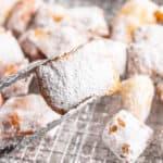 A pair of tongs holding a beignet over the baking sheet.