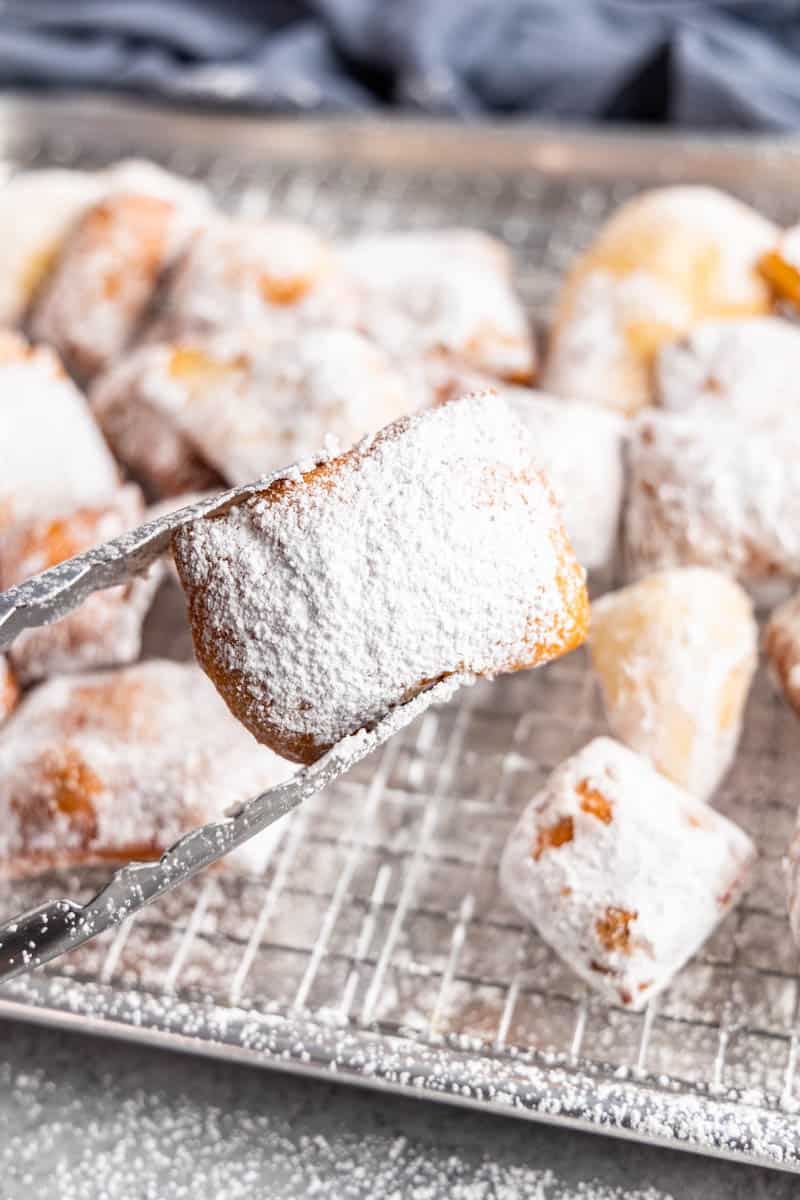 A pair of tongs holding a beignet over the baking sheet.