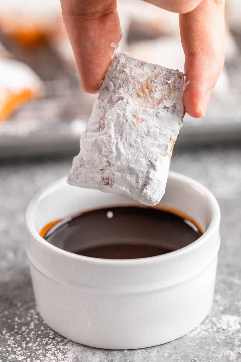 A hand dipping a beignet into chocolate sauce.