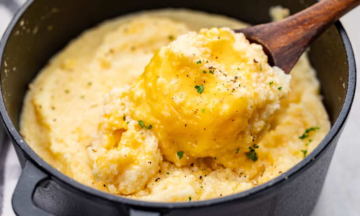 A wooden spoon dipped into a pot filled with cheesy grits.