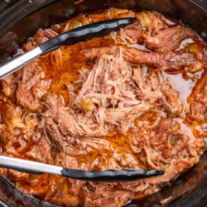 A pair of tongs in a slow cooker filled with sweet pork.